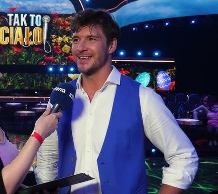 Mikołaj Roznerski na scenie teatralnej, oświetlony reflektorami, symbolizujący jego karierę aktorską i różnorodność ról.