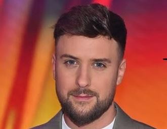 Aleksander Sikora, prezenter telewizyjny, na scenie podczas wydarzenia, w eleganckim garniturze, z uśmiechem, symbolizujący dynamikę i profesjonalizm.