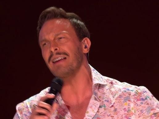 Alt: Sławek Uniatowski śpiewający na scenie podczas koncertu, z zespołem w tle, oświetlony teatralnymi światłami.