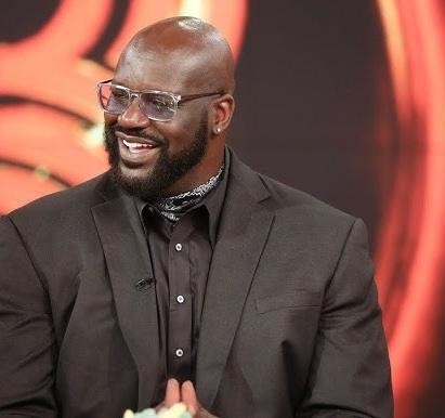 Portret Shaquille'a O'Neala w stroju biznesowym, uśmiechniętego, z rozmytym tłem sugerującym środowisko akademickie lub biznesowe.