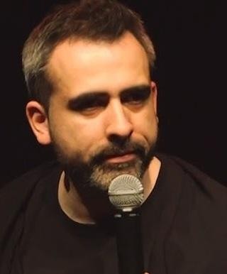 Abelard Giza na scenie podczas występu stand-upowego, dynamicznie gestykulujący z mikrofonem, z wciągającym, humorystycznym wyrazem twarzy, nawiązujący kontakt z publicznością.
