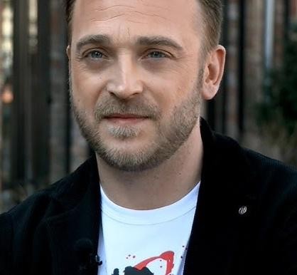 Mateusz Gessler, polski restaurator i osobowość telewizyjna, uśmiecha się w nowoczesnej kuchni, trzymając nóż szefa kuchni i pięknie podane danie.