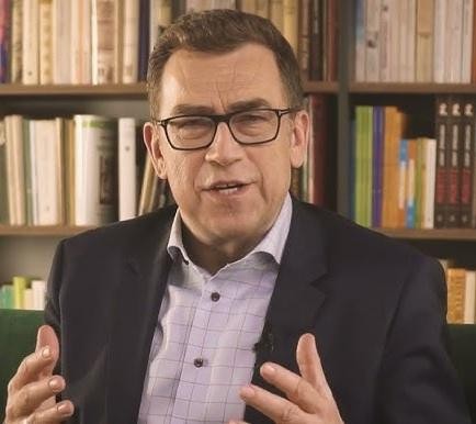 Maciej Orłoś jako prezenter telewizyjny, z charakterystycznym gestem, w nowoczesnym studiu informacyjnym.