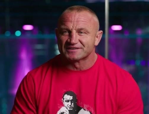 Alt: Mariusz Pudzianowski w oktagonie MMA, z zaciętą miną podczas walki, prezentujący swoją muskulaturę i skupienie, z subtelnym logo KSW w tle.