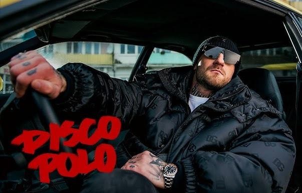 Kizo (Patryk Woziński) w koszulce firmowej, prezentujący jeden ze swoich produktów spożywczych, batona Kizzers lub pizzę Sukcesiliana.